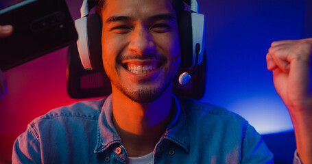 Happy asia man gamer wear headphone competition play video game online with smartphone colorful neon light in living room at night modern house. Esport streaming game online, Home quarantine activity. © tirachard
