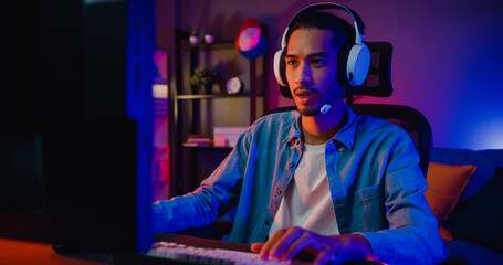 Happy asia guy gamer wear headphone participation play video game colorful neon lights computer in living room at night modern house. Esport streaming game online, Home quarantine activity concept. - obrazy, fototapety, plakaty