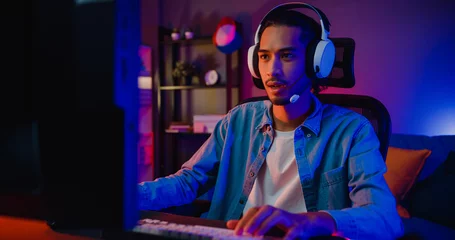 Fotobehang Gaming Happy asia guy gamer wear headphone participation play video game colorful neon lights computer in living room at night modern house. Esport streaming game online, Home quarantine activity concept.  © tirachard