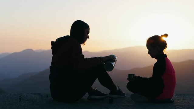 Young mother and 9 years daughter are spending time together at sunset on observation deck. They drinks tea with beautiful mountains view. Silhouette 4k footage.