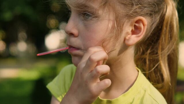 Close-up of a beautiful girl eating ice cream with a red spoon and fixing her disheveled hair with her hand. A child in a city park eating ice cream. Carefree happy childhood