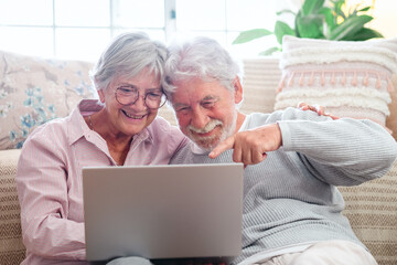 Cheerful caucasian senior couple sitting on the floor at home using laptop, modern retired elderly people surfing the net with computer