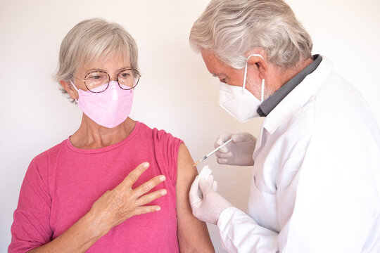 Doctor giving injection to senior caucasian woman, female receiving fourth booster of covid 19 vaccine, people wearing mask