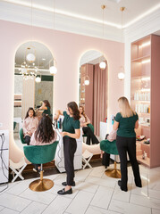 Two female hairstylists curling client hair and doing hairstyle in modern beauty salon. Young women sitting in chairs in front of mirrors while hairdressers styling clients hair.