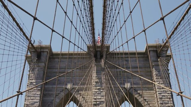The Brooklyn Bridge with an American Flag on the top during a sunny day with blue skies, tilt shot.