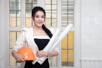 Female architect or engineer holding blueprints and work helmet near office windows.