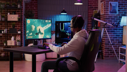 African american man playing fast paced multiplayer space shooter using pc gaming setup having a good time in home living room. Gamer streaming online simulation game while talking in headset.