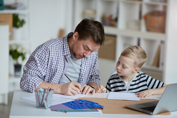 Serious young father sitting at table with son and drawing on paper while assisting son with homework