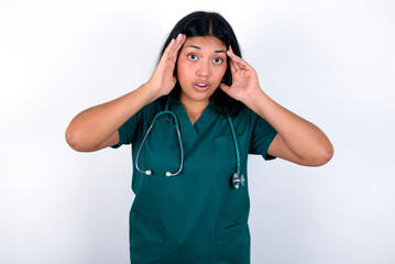 Doctor hispanic woman wearing surgeon uniform over white wall with scared expression, keeps hands on head, jaw dropped, has terrific expression. Omg concept