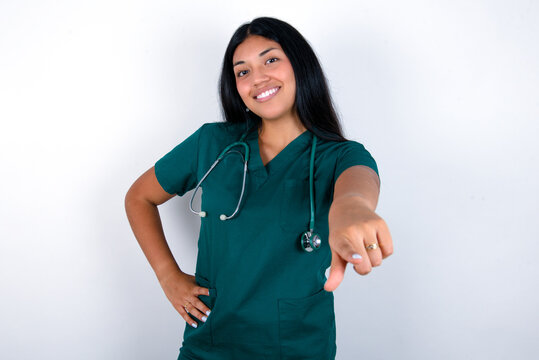 Doctor hispanic woman wearing surgeon uniform over white wall pointing at camera with a satisfied, confident, friendly smile, choosing you