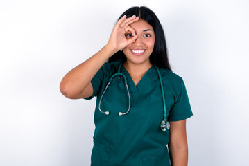 Doctor hispanic woman wearing surgeon uniform over white wall doing ok gesture with hand smiling, eye looking through fingers with happy face.