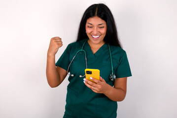 Doctor hispanic woman wearing surgeon uniform over white wall holding in hands cell and rising his fist up being excited after reading good news.