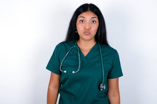 Shot of pleasant looking Doctor hispanic woman wearing surgeon uniform over white wall, pouts lips, looks at camera, Human facial expressions