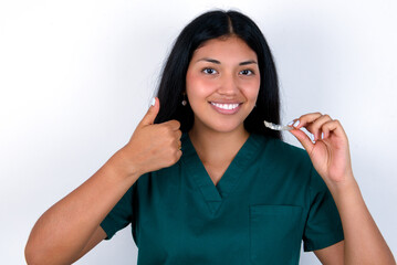 Doctor hispanic woman wearing surgeon uniform over white wall holding an invisible braces aligner...