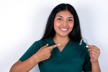 Doctor hispanic woman wearing surgeon uniform over white wall holding an invisible aligner and pointing at it. Dental healthcare and confidence concept.