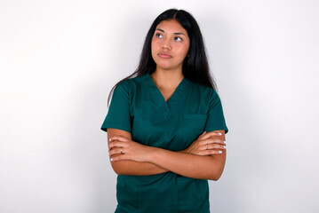 Charming thoughtful Doctor hispanic woman wearing surgeon uniform over white wall stands with arms...