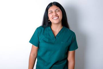 Positive Doctor hispanic woman wearing surgeon uniform over white wall with overjoyed expression closes eyes and laughs shows white perfect teeth