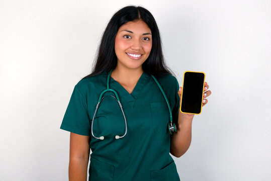 Smiling Doctor hispanic woman wearing surgeon uniform over white wall Mock up copy space. Hold mobile phone with blank empty screen