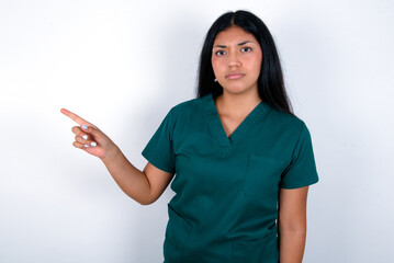 Serious Doctor hispanic woman wearing surgeon uniform over white background smirks face points away on copy space shows something unpleasant. Look at this advertisement. Big price concept