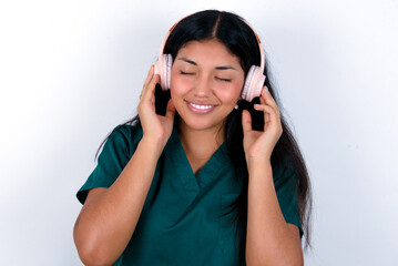Pleased Doctor hispanic woman wearing surgeon uniform over white background enjoys listening pleasant melody keeps hands on stereo headphones closes eyes. Spending free time with music