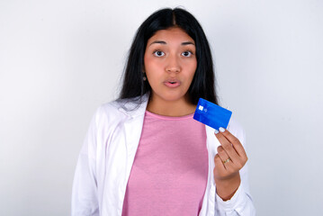 Photo of Young hispanic doctor girl wearing coat over white background amazed shocked hold credit card payment