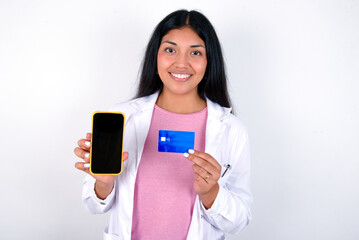 Photo of adorable Young hispanic doctor girl wearing coat over white background holding credit card and Smartphone. Reserved for online purchases