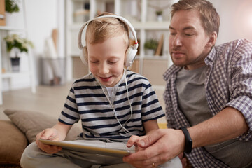 Positive cute blond-haired boy in wired headphones using learning app on tablet together with father at home
