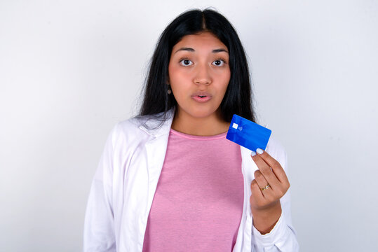 Photo of Young hispanic doctor girl wearing coat over white background amazed shocked hold credit card payment - Powered by Adobe