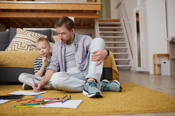 Fototapeta premium Serious creative young father in casual outfit sitting on carpet and drawing with colorful pencils together with son