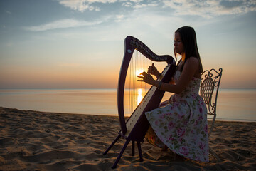 A girl in a flower dress plays on a Celtic harp by the sea at sunset