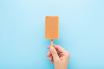 Young adult woman hand holding light brown caramel ice cream with stick on light blue table background. Pastel color. Closeup. Top down view.