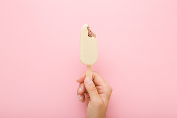 Young adult woman hand holding chocolate ice cream with white vanilla glaze on stick. Light pink table background. Pastel color. Bitten food. Closeup. Cold sweet snack in summer. Top down view.