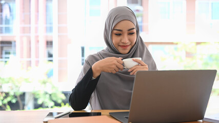 Pleased muslim lady in hijab holding cup of coffee and reading business email or financial information on laptop