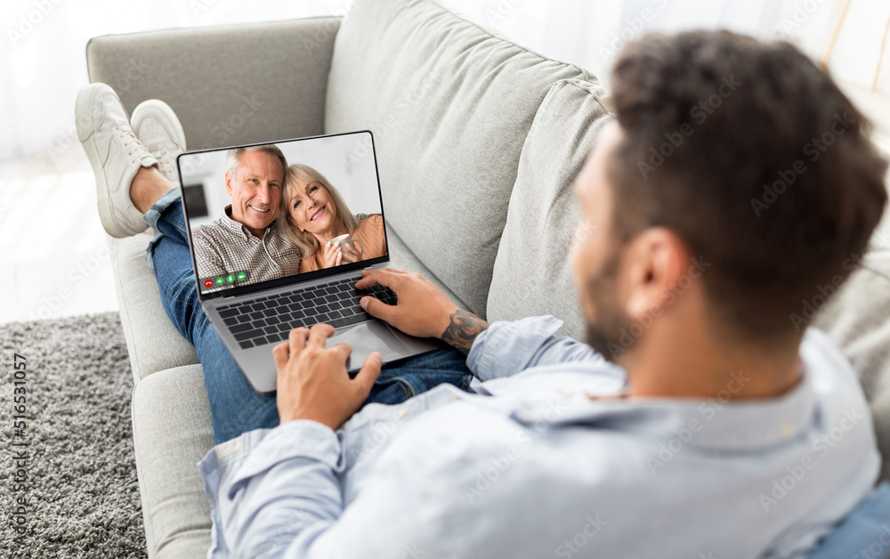 Poster man making video call on laptop with senior parents while relaxing on couch