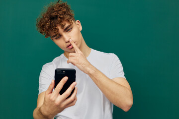 a funny man with curly hair stands on a green background in a gray T-shirt and holds his finger near his nose while holding the phone in his hand