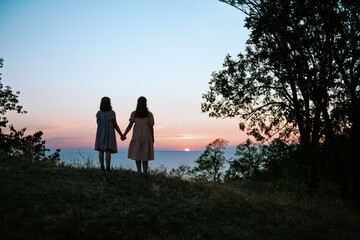 Silhouette of two teen sisters from behind holding hands and watching beautiful summer sunset over the sea 