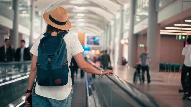 A cinematic handheld back shot of a woman with a backpack and a hat standing on a moving walkway of an airport.