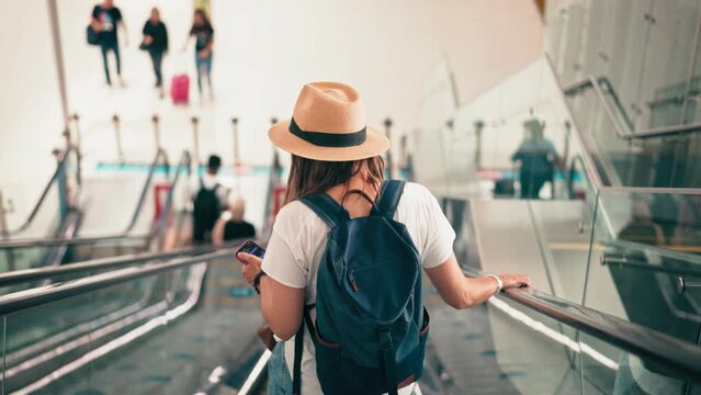 A cinematic hand-held back shot of a woman with a backpack and a hat standing on an escalator going down in an airport or shopping mall.