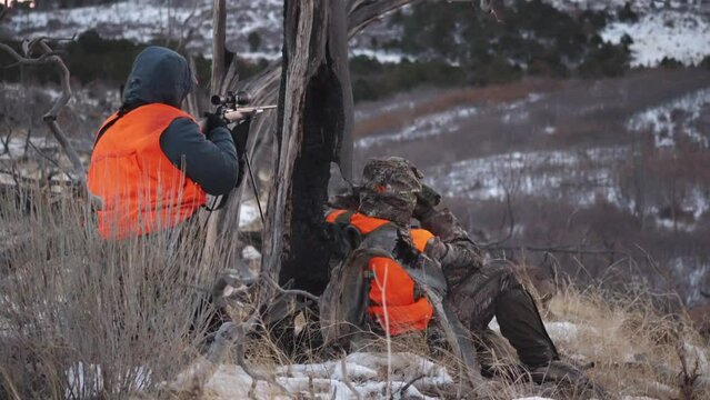 Two hunters sit scouting on a cold mountain side while looking in the distance and one prepares to shoot his gun, static shot