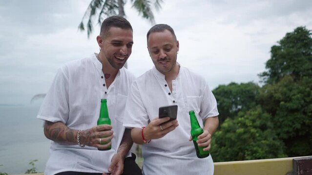 Two happy men, gay couple honeymooners holding beer and watching phone pictures during summer vacation in a tropical Island