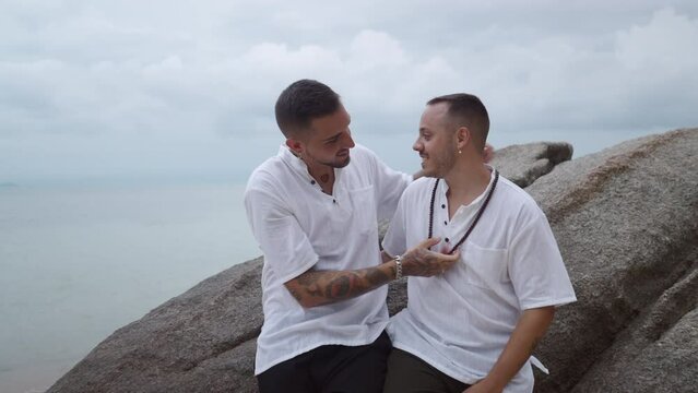 Gay couple on honeymoon. Man wearing a handmade necklace on the neck of his partner and kiss him sitting on the big rock by the sea 