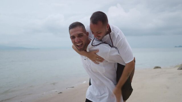 Cheerful gay man giving piggyback ride to his partner on a beach  - Powered by Adobe