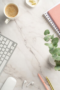 Business concept. Top view vertical photo of workspace keyboard cup of coffee pink notepads pens gold binder clips computer mouse wireless earbuds and eucalyptus on white marble background