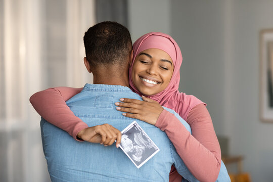 Pregnancy News. Pregnant Black Muslim Woman Holding Sonography Picture And Hugging Husband