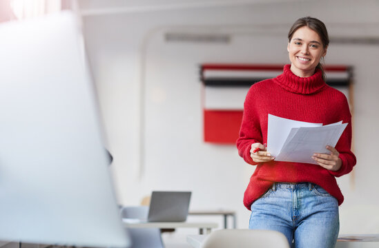 Portrait Of Happy Attractive Young Woman In Red Warm Sweater Standing In Modern Office And Viewing Design Project Papers