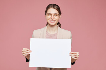 Fototapeta premium Portrait of happy beautiful intelligent young woman in eyeglasses standing against pink background and holding blank poster, advertising message concept