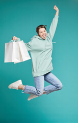 Portrait of positive excited hipster girl in hoodie jumping with purchases after shopping sale,...