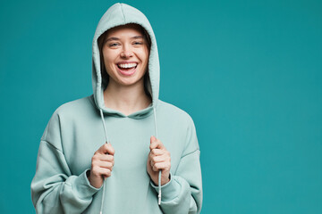 Portrait of happy beautiful girl in teal hoodie pulling strings down and laughing cheerfully...