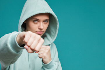 Portrait of serious female fighter in teal hoodie punching at camera while preparing for fight, isolated background