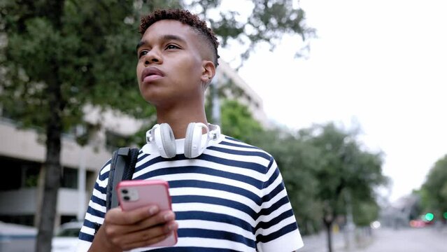 Young hispanic latin man in striped shirt with headphones holding smart mobile phone standing outdoor looking for address with smartphone app in city street